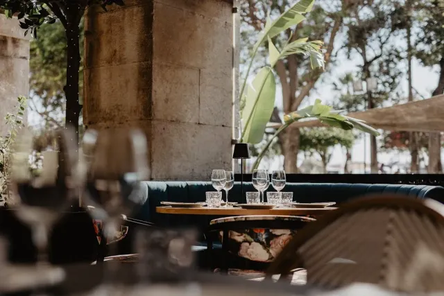 A table set with wine glasses facing a blue banquette, stone columns and a broad leaved plant