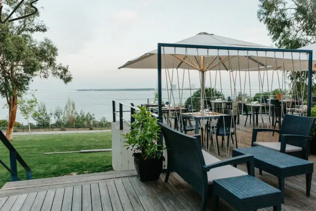 A wooden deck with a large parasol and rattan armchairs, a lawn and a wide bay behind