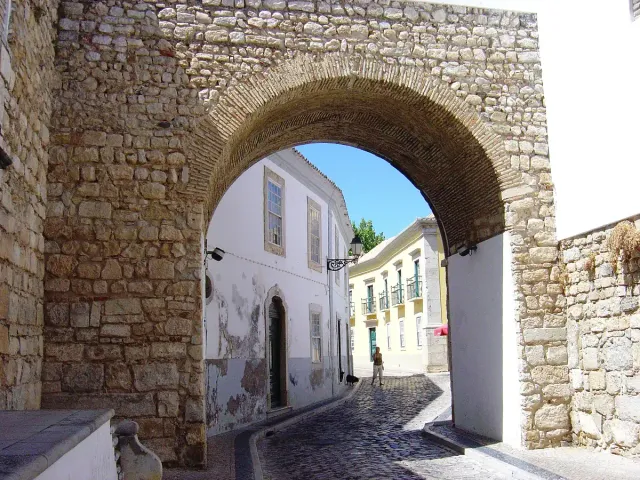 An arched stone gate in a wall leading to a cobbled street of white houses