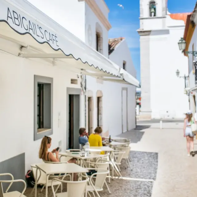 A white alley with a scalloped awning, pale metal tables and chairs and a bell tower behind