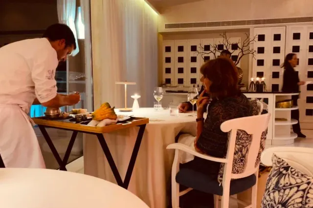 A cook plating a dish on a trolley beside a seated guest in a softly lit room