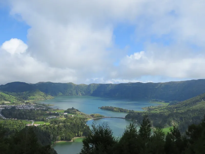 A wide lake inside a green volcanic crater, with a small village and a bridge crossing the water