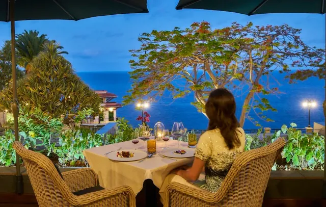 A woman seated at a set table under a parasol facing a flowering tree and the sea in the evening