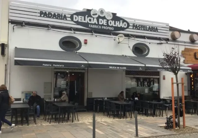 A bakery front with a large sign, dark awnings and metal tables on the pavement