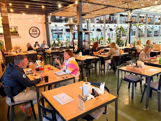 A covered hall with wooden tables, string lights and diners along the glass partition