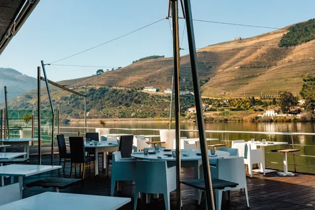 A wooden terrace with white tables above a river, and terraced hills across it