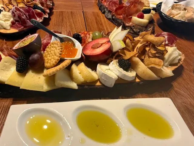 A wooden board loaded with cheeses, cured meats, berries and crackers beside three dishes of olive oil