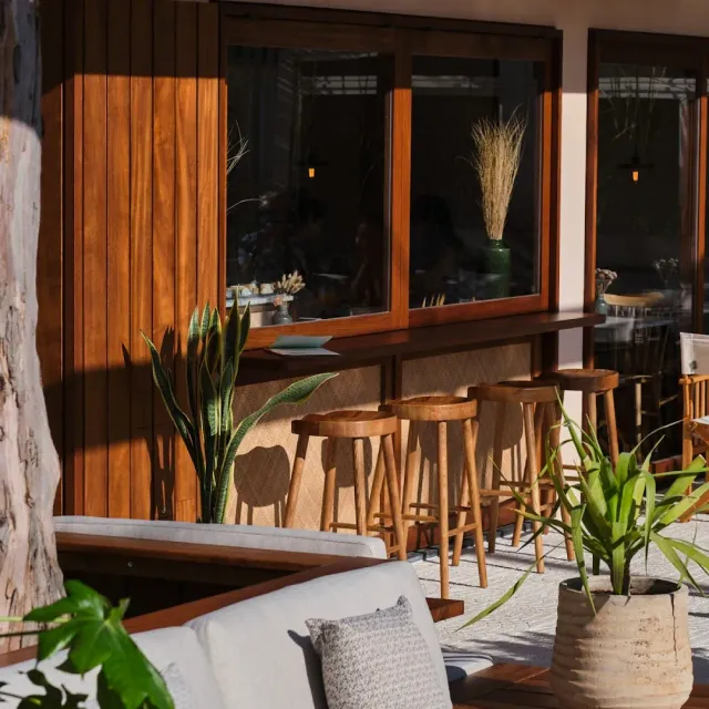 A wooden front with a bar and high stools, potted plants, a white sofa and dappled sunlight