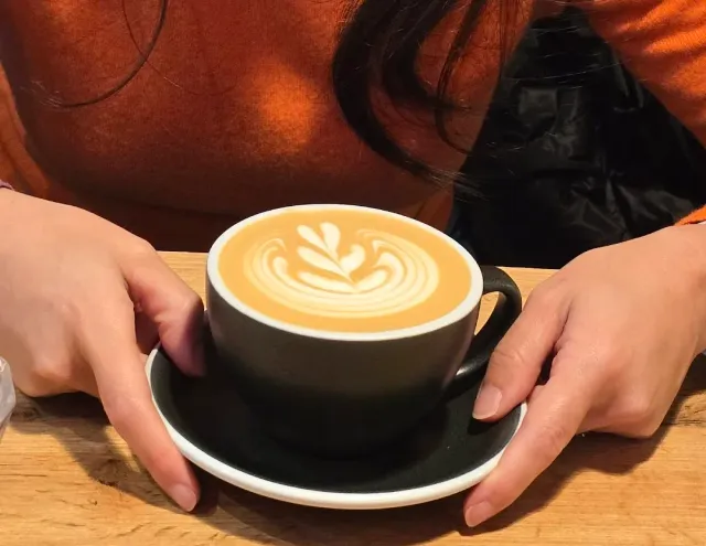 Hands holding a black cup with a leaf drawn in the foam, on a saucer and a wooden table