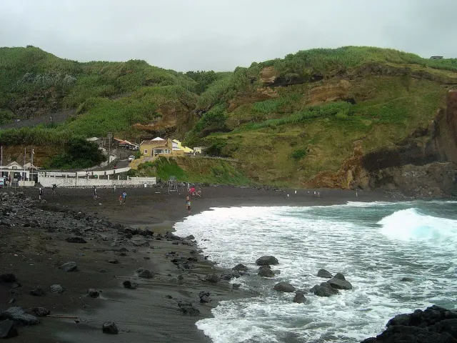 A dark sand beach with sea rocks and white waves below a green slope