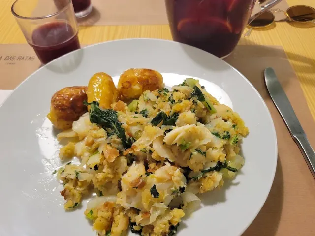 A plate of shredded cod with greens, fried potatoes and glasses of red wine behind