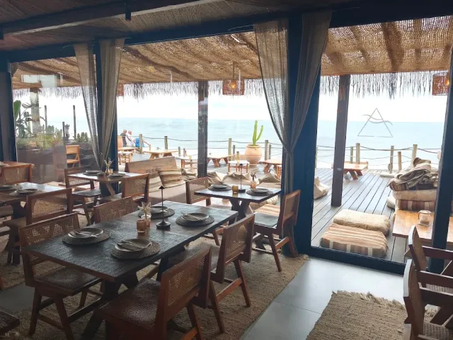 A space with a straw roof, rattan chairs and wooden tables opening onto a deck facing the sea