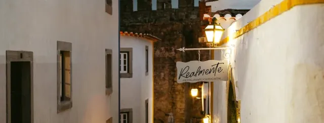 A narrow alley between white houses with a hanging sign and a lit lantern at dusk