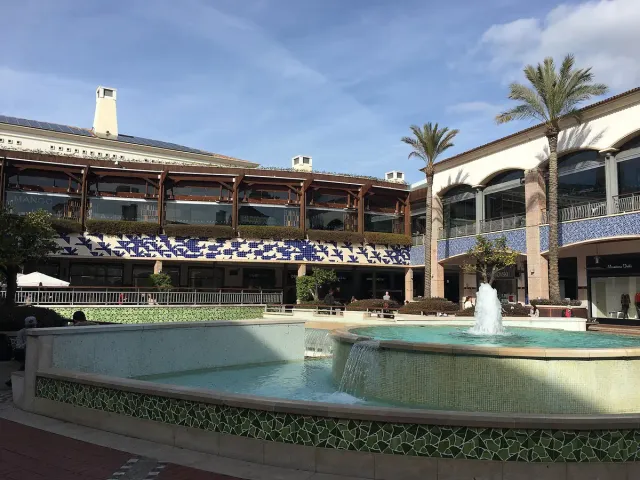 A fountain in a pool before open shop galleries and tall palms