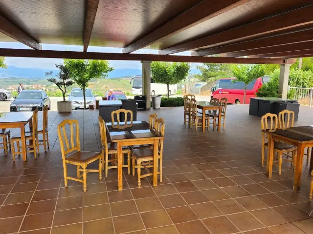 A terrace covered by a wooden pergola with tables and chairs, and cars and hills behind it