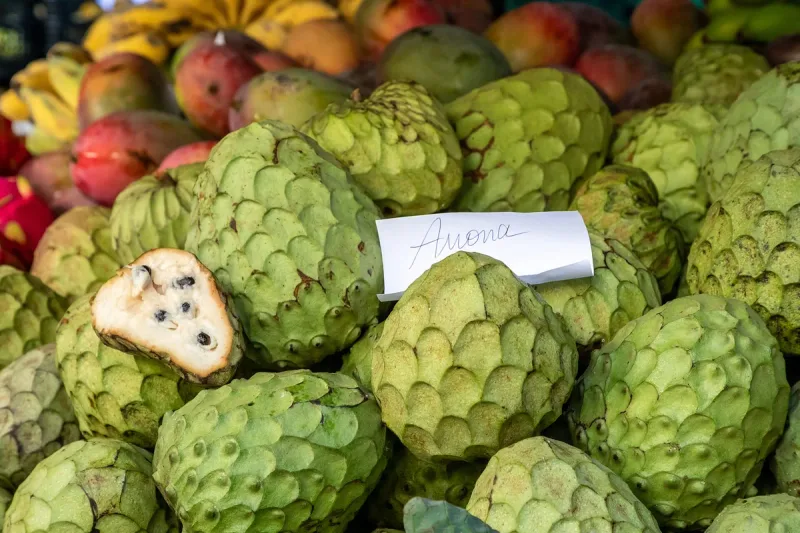 A pile of green custard apples on a stall, one cut open to show white flesh and black seeds