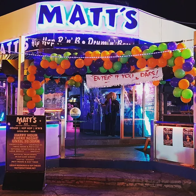 A lit bar entrance at night, decorated with an arch of orange, purple and green balloons