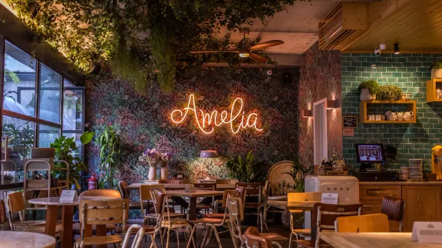 A space with floral wallpaper, an orange neon sign, hanging greenery and small wooden tables