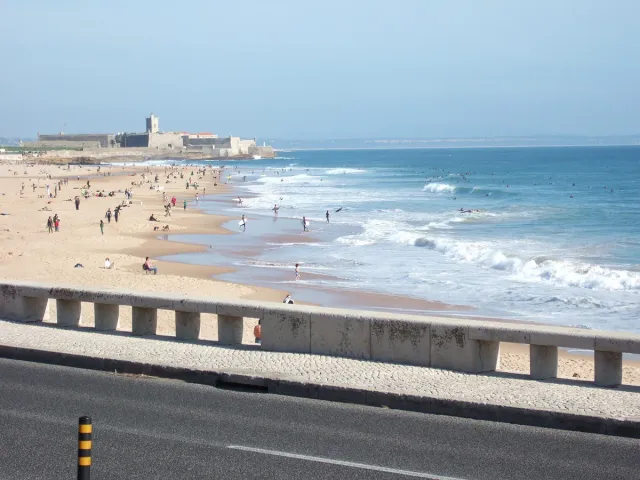 A long sandy beach with bathers, white waves and a fort at the far end
